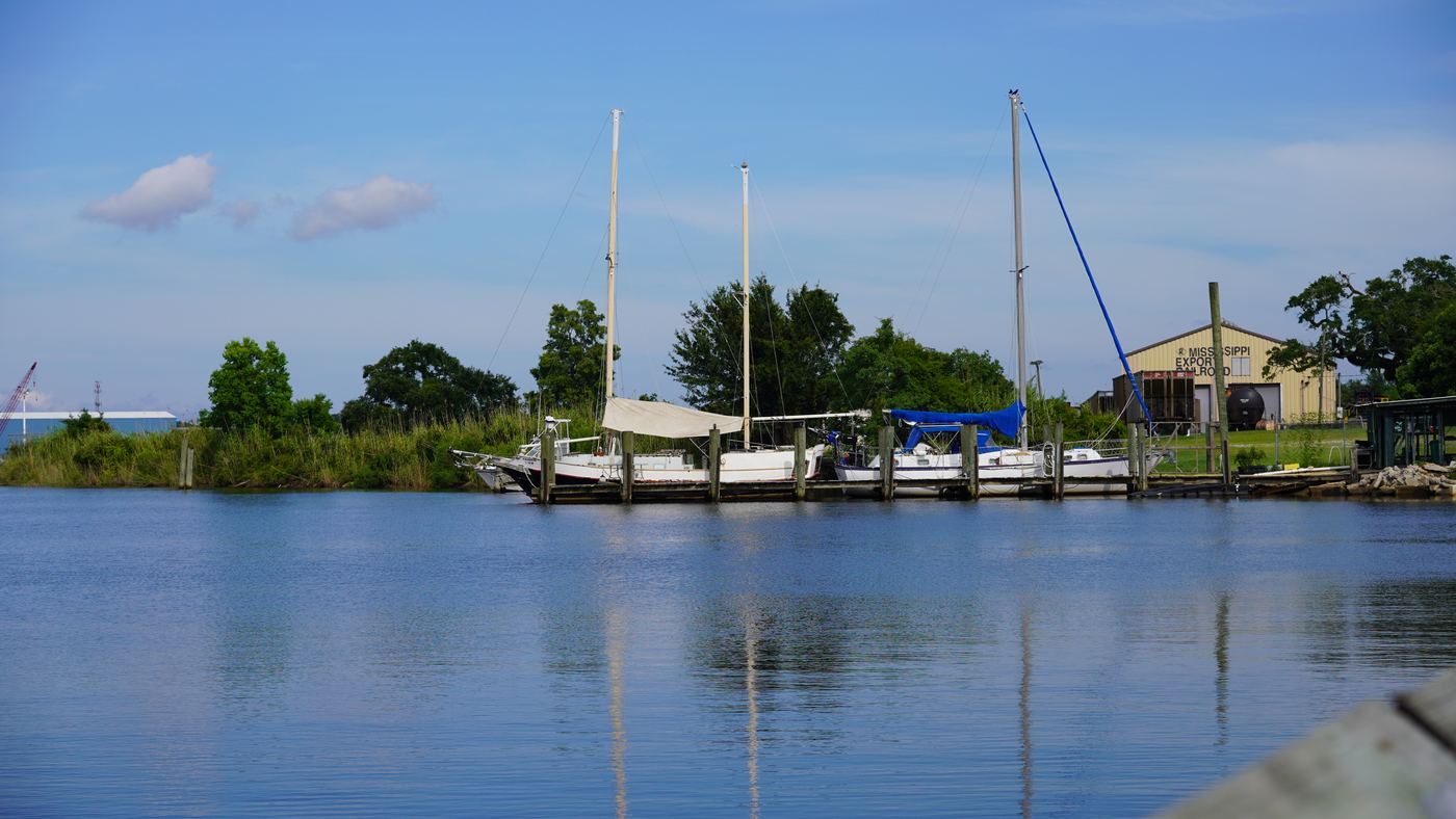 Sailboats moored at a quiet marina on the river in Moss Point, with a boathouse and calm reflecting water.