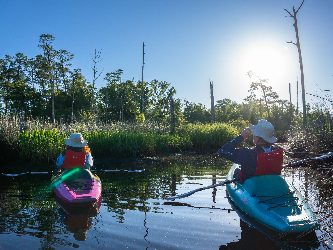 Two kayakers paddle a narrow marsh channel near the Pascagoula River Audubon Center, the morning sun low over the grasses.
