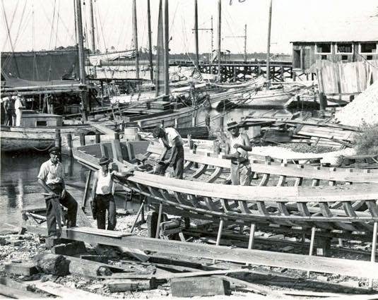 Workers shape the wooden hulls of boats under construction beside the water at Moss Point.