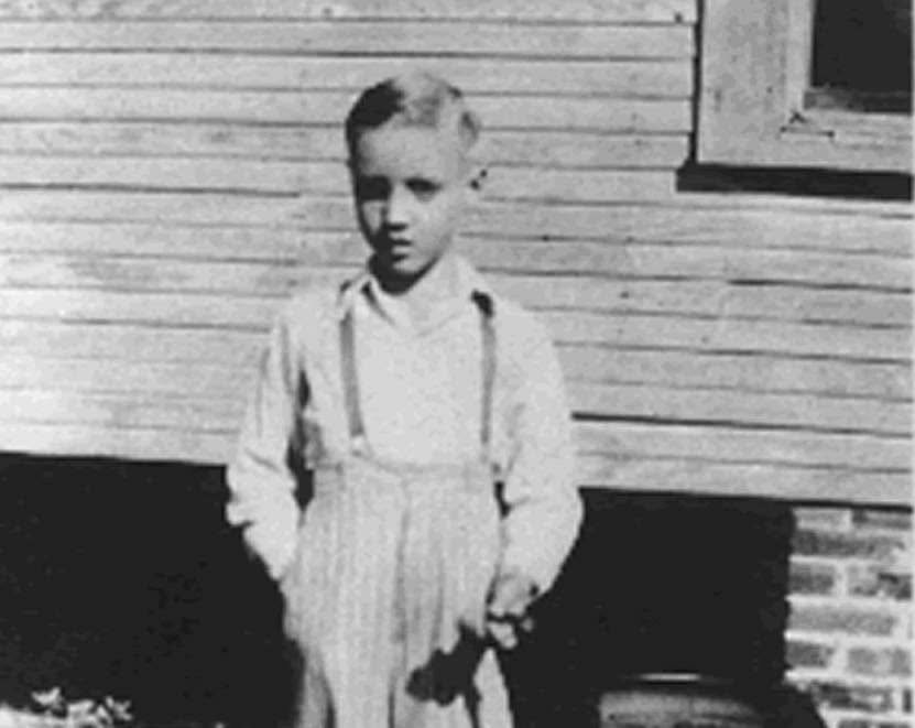 A young boy in suspenders stands against a clapboard wall in the early 1940s, said to be Elvis Presley.