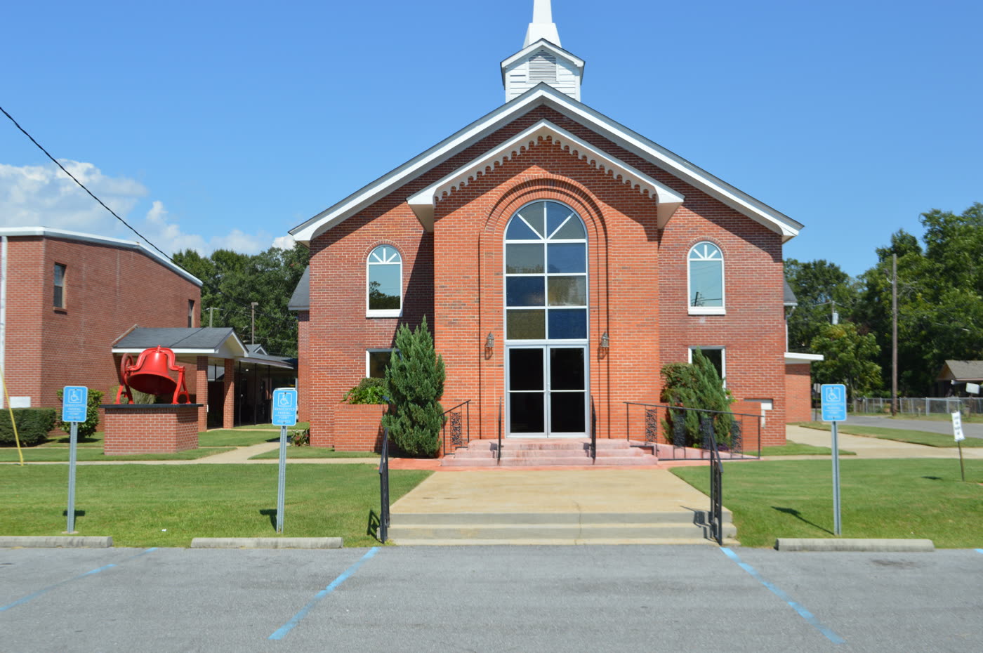 The red brick sanctuary of First Missionary Baptist Church in Moss Point, with a white steeple, arched windows, and the church's red bell displayed out front.