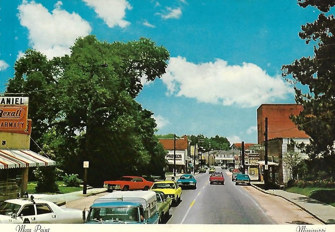 A color photograph of Main Street in Moss Point around 1970, with period cars parked along the road and a Daniel Rexall pharmacy sign on the left.