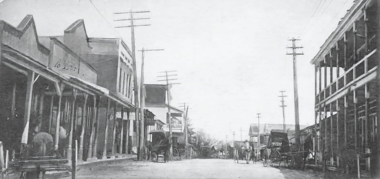 A wide unpaved Main Street lined with two story wood front stores and utility poles in Moss Point around 1898, with horse drawn wagons.