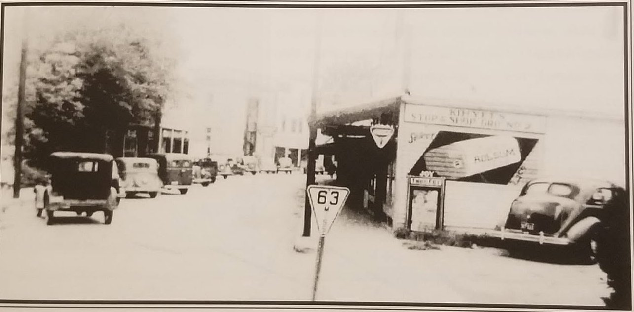 Parked cars line a paved 1930s Main Street in downtown Moss Point with a Highway 63 sign and storefronts.