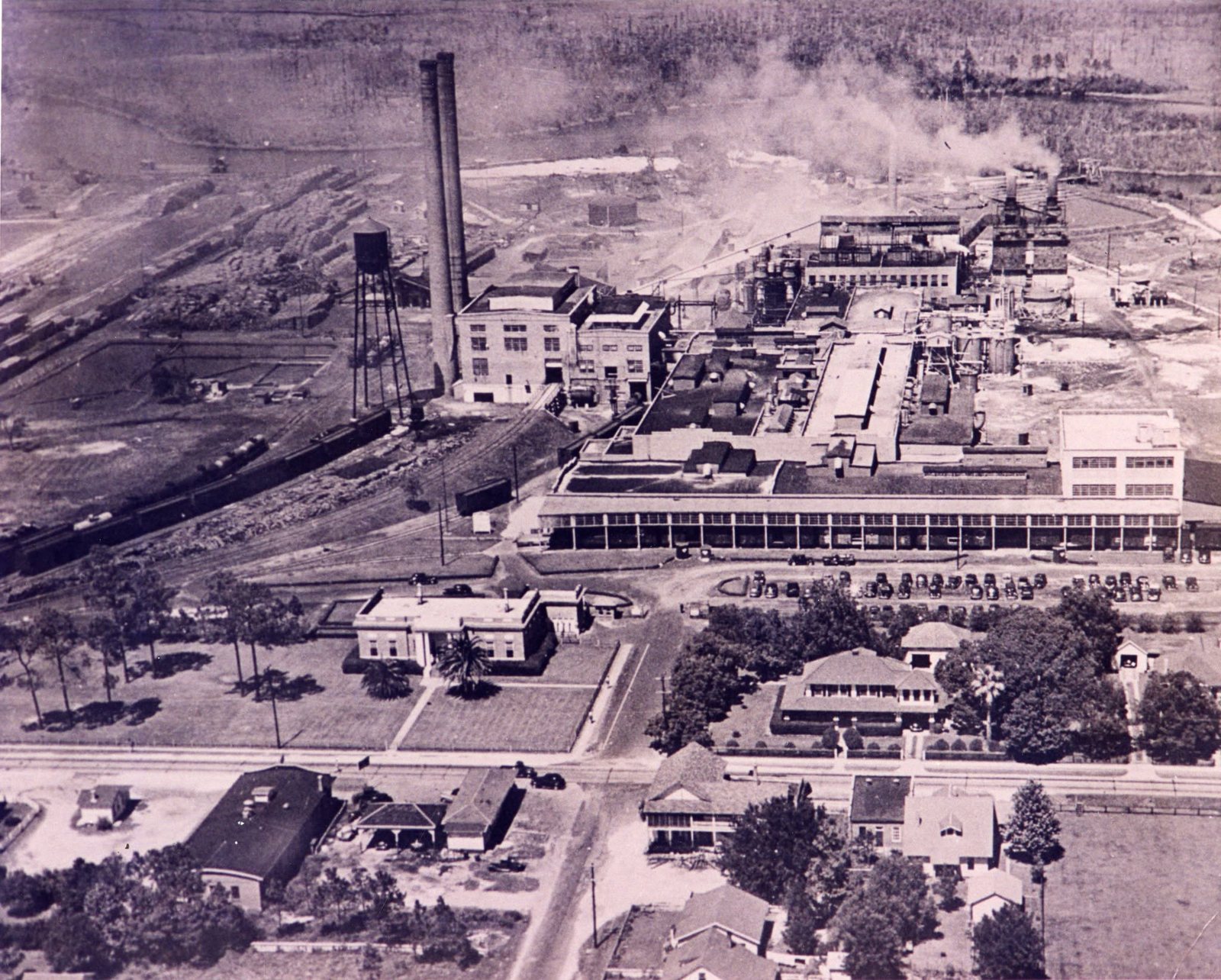 An aerial view of the Moss Point paper mill, its smokestack and long buildings beside the river, with homes in the foreground.