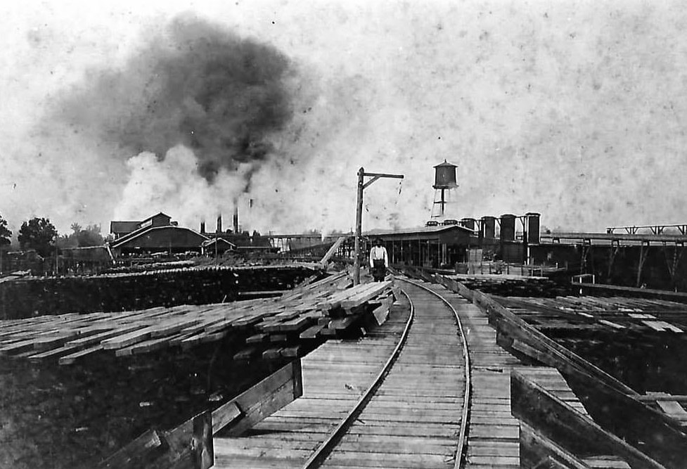 A Moss Point sawmill with a wooden tramway track running out through acres of stacked drying lumber, smoke rising from the mill and a water tower beyond.