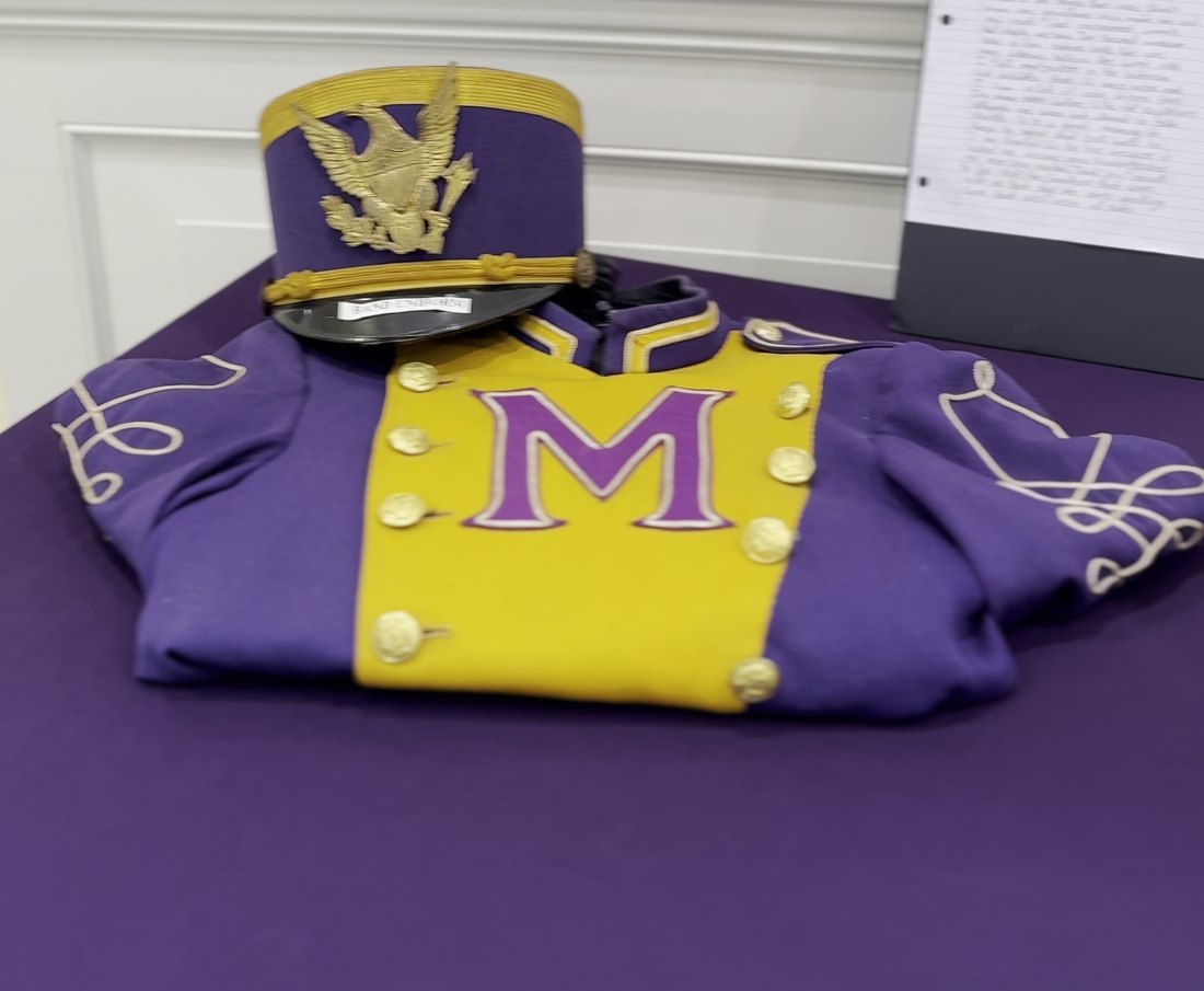 A Magnolia High School Monarch marching band uniform in purple and gold with a large letter M, beside its plumed shako hat with a gold eagle crest.