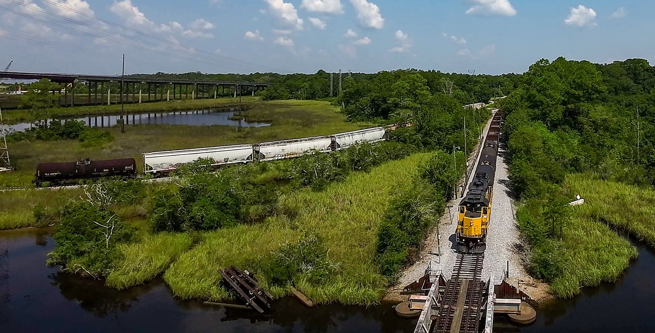 A yellow Mississippi Export Railroad locomotive hauls tank cars across a marsh trestle at Moss Point, the highway bridge beyond.