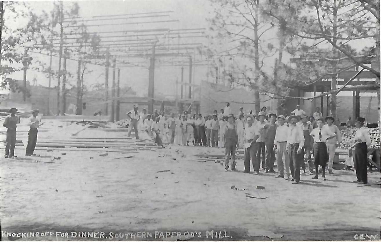 A large crowd of workers in work clothes pose in the yard of the Southern Paper Company mill as its steel framework rises behind them.