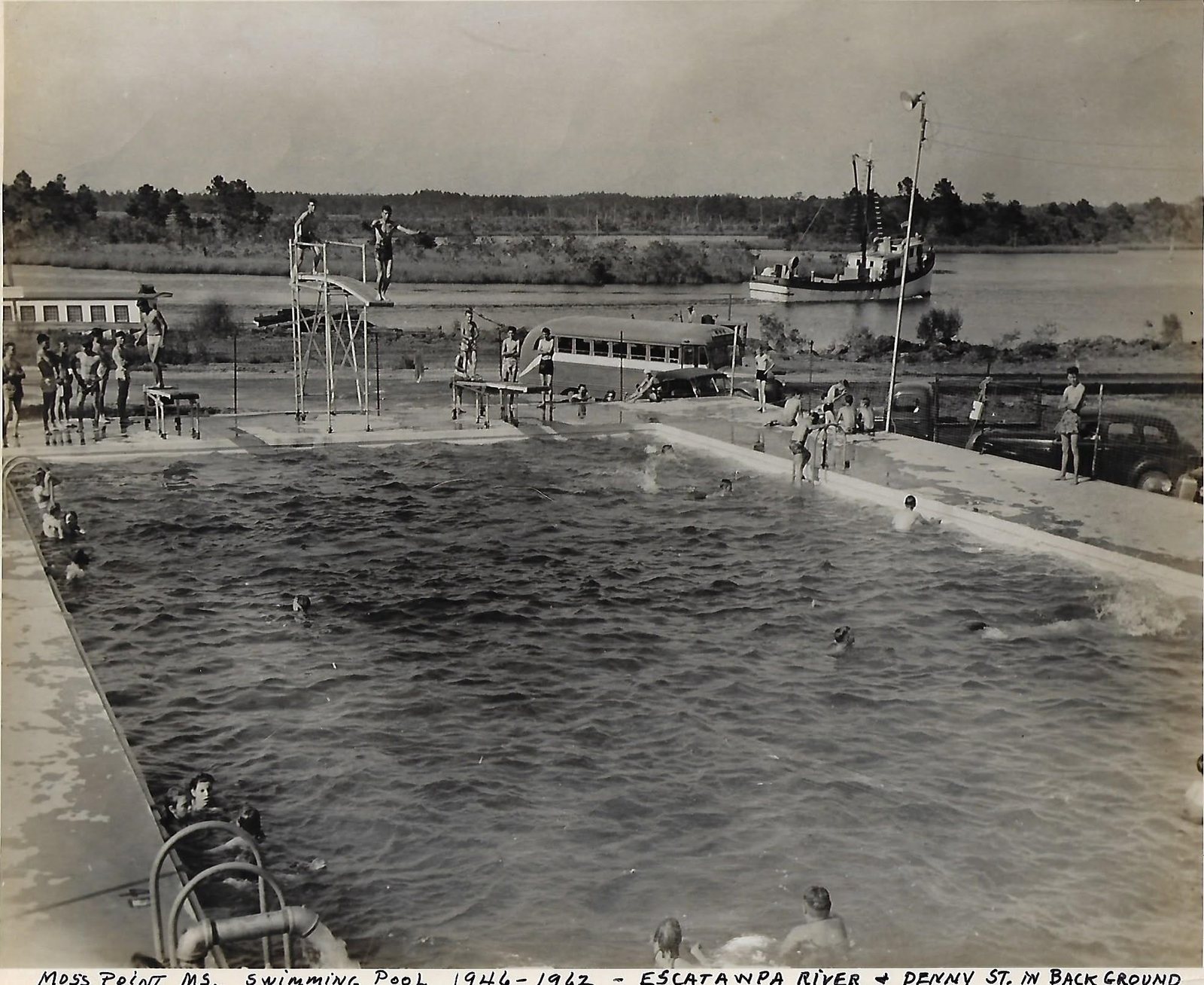 Swimmers fill an outdoor municipal pool with diving boards while a shrimp boat passes on the Escatawpa River behind, in the mid 1940s.