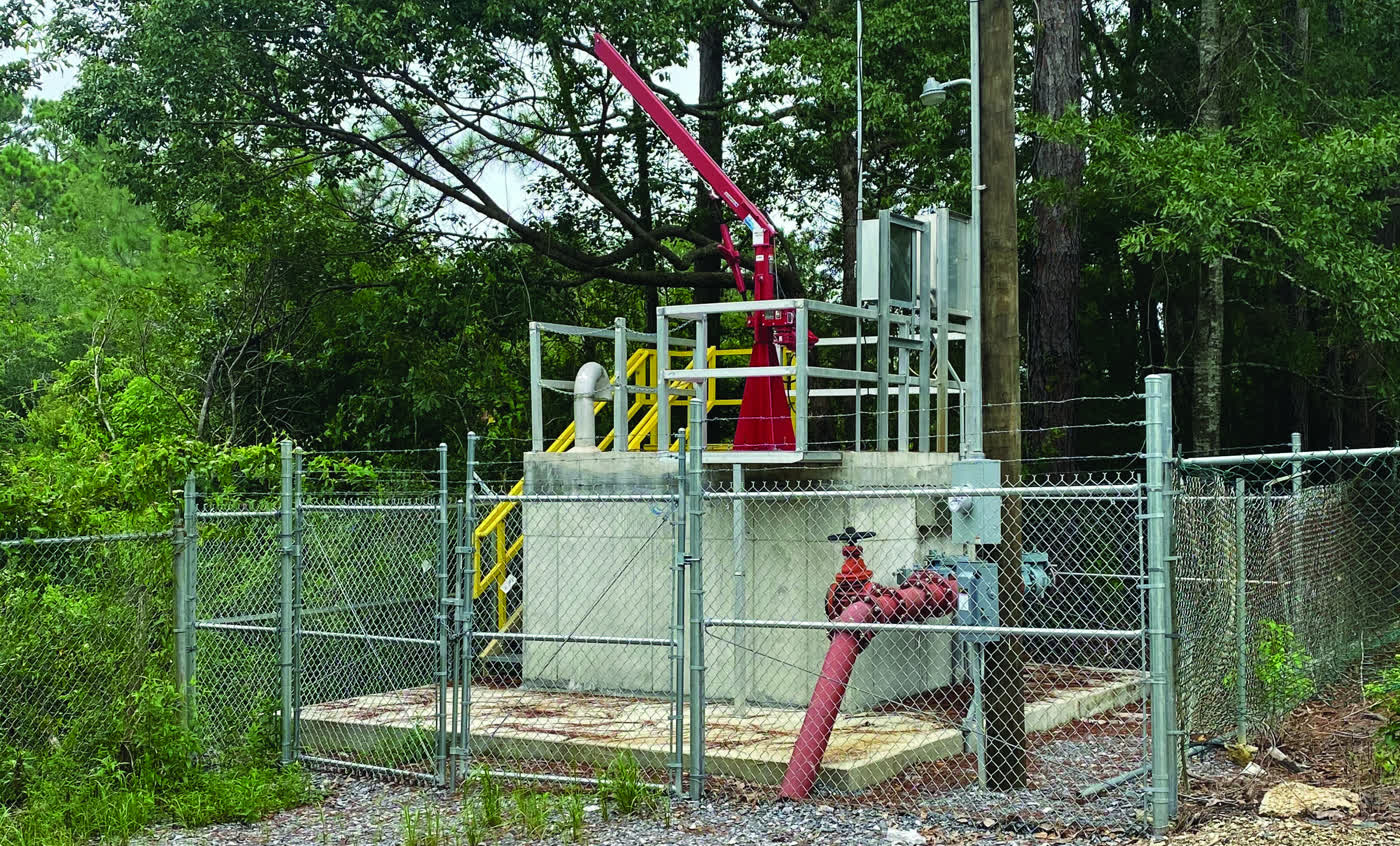 A rehabilitated sewer pump station in Moss Point, raised on a concrete platform above flood level, with a red davit crane and new controls behind a chain link fence.