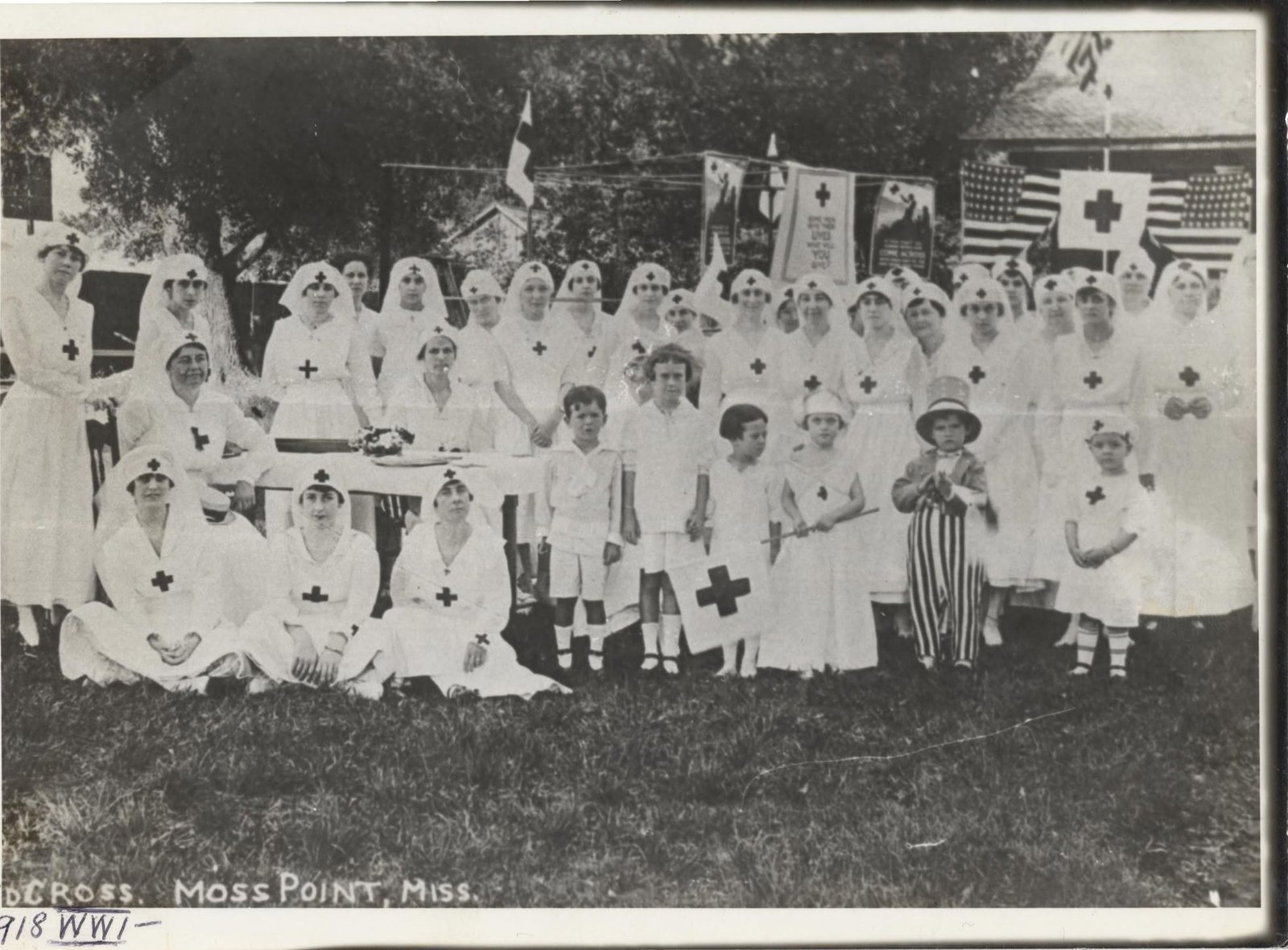 Several dozen women and children in white Red Cross uniforms pose beneath American flags and Red Cross banners in Moss Point in 1918.