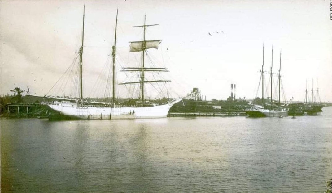 A large square rigged sailing ship and several schooners moored at the Moss Point waterfront with mill smokestacks behind, early 1900s.