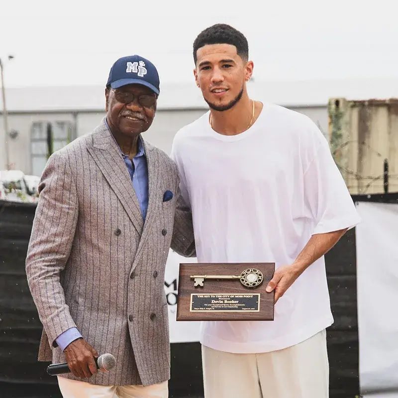 Mayor Billy Knight in a suit presents NBA All Star Devin Booker the key to the city.