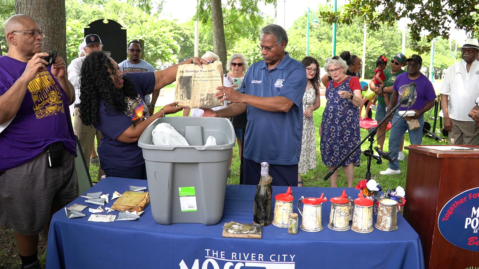 Two people hold up a yellowed Mississippi Press Register front page recovered from the capsule, with steins and papers on a table draped in a City of Moss Point banner as the community looks on.