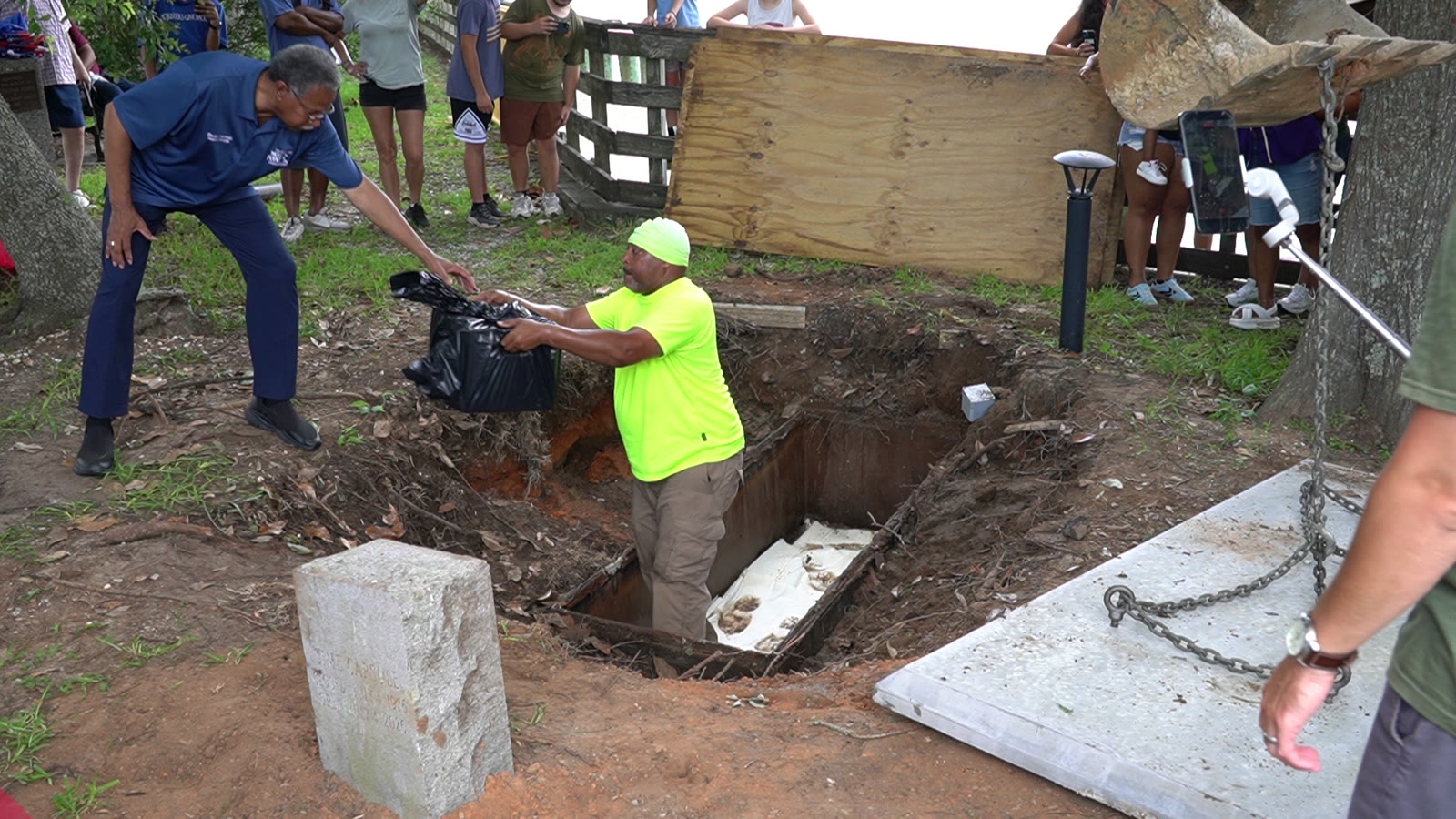 Alderman David Chapman reaches down to take a black bag of the recovered 1976 time capsule contents from a Moss Point public works worker standing in the freshly dug riverfront pit, as a crowd watches.