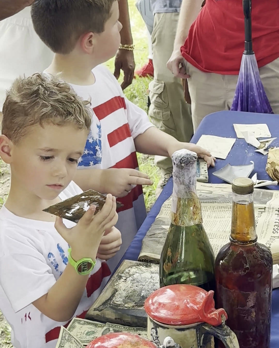 Two boys in patriotic Fourth of July shirts examine keepsakes from the 1976 time capsule laid out on a table, including old glass bottles, a stein with a red lid, a dollar bill, and a yellowed newspaper.