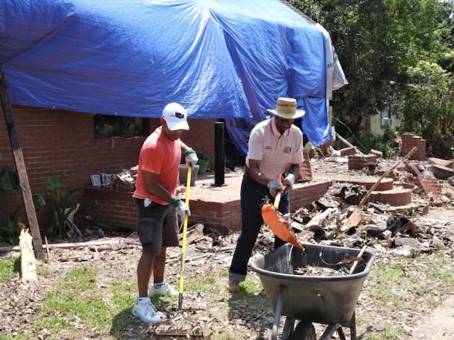 Mayor Billy Knight Sr. in a straw hat shovels storm debris into a wheelbarrow alongside Dr. Washington, in front of a tarped, tornado-damaged brick home in Moss Point.