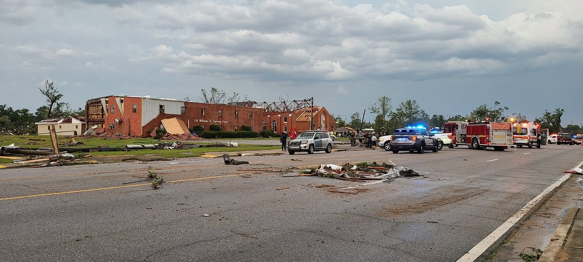 A wide view of tornado damage in Moss Point: the J.W. Williams, Sr. Learning Center with its roof torn off, debris across the highway, and police, fire, and ambulance crews on the scene.