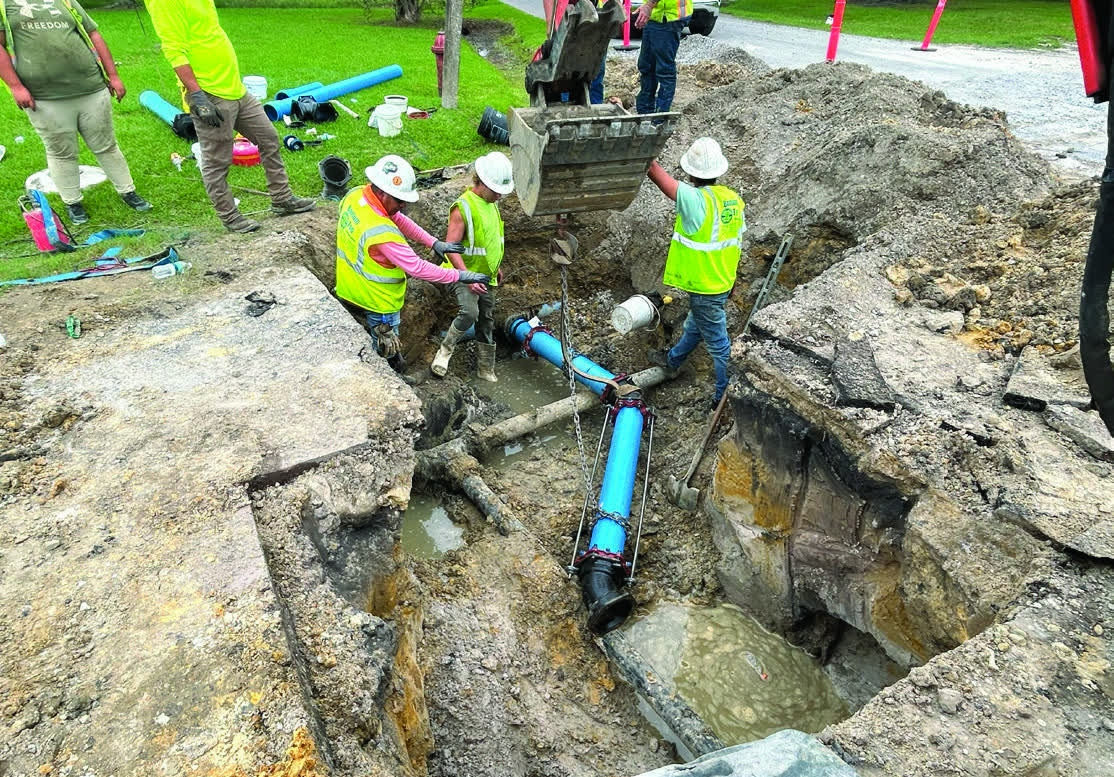 A work crew in hard hats and safety vests lowers new blue PVC water main into an open trench on Kimberly Drive in Moss Point.