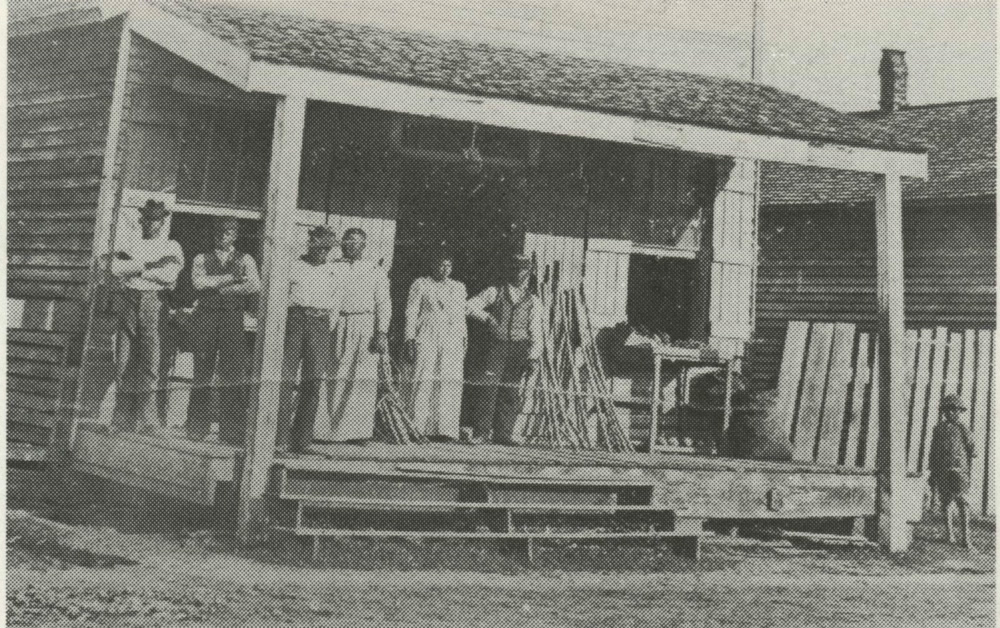 Ada Williams stands in the doorway of her wooden store, its deep porch stacked with goods, customers on the porch and a child at the right.