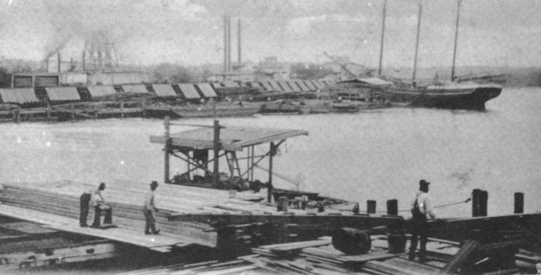 Workers stand on stacked sawn lumber on a wharf at the L.N. Dantzler mill, a loading rig behind them and a schooner moored on the river.