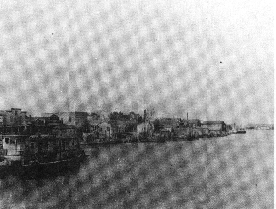 The Moss Point waterfront around 1900 seen from the river, a riverboat at the left and wooden stores and docks lining the bank.