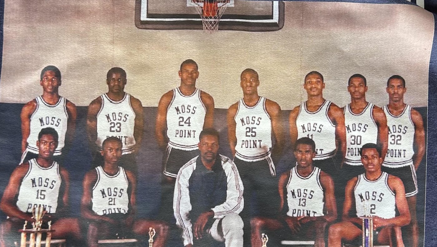 Coach Arthur Haynes kneeling with a basketball, surrounded by eleven Tigers in Moss Point jerseys and a row of trophies, from a printed 1987 team photograph.