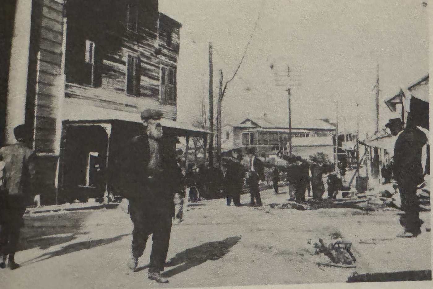A crowd of men on a dirt Main Street near a two story wooden building during a fire, storefronts and poles beyond.