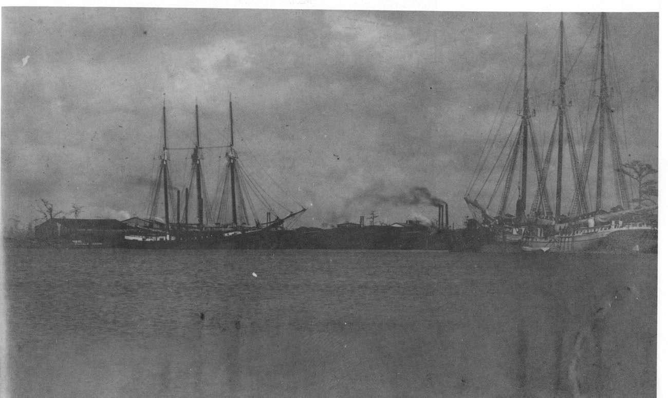 Two tall lumber schooners at anchor on McInnis Lake, the McIntosh Lumber Company sawmill smoking on the far shore.