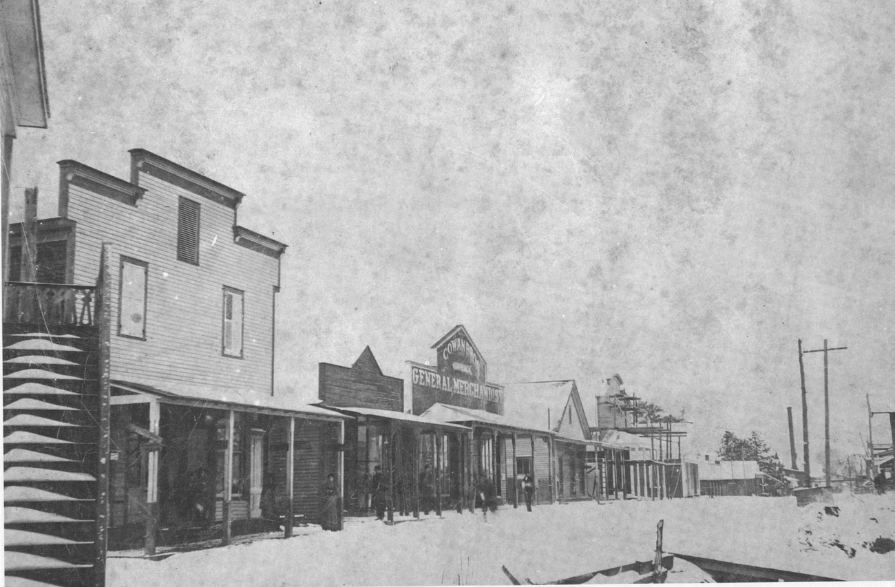 Snow blankets Main Street in Moss Point around 1920, wooden false-front stores including Cowan Brothers General Merchandise lining the street, figures standing on the boardwalk.