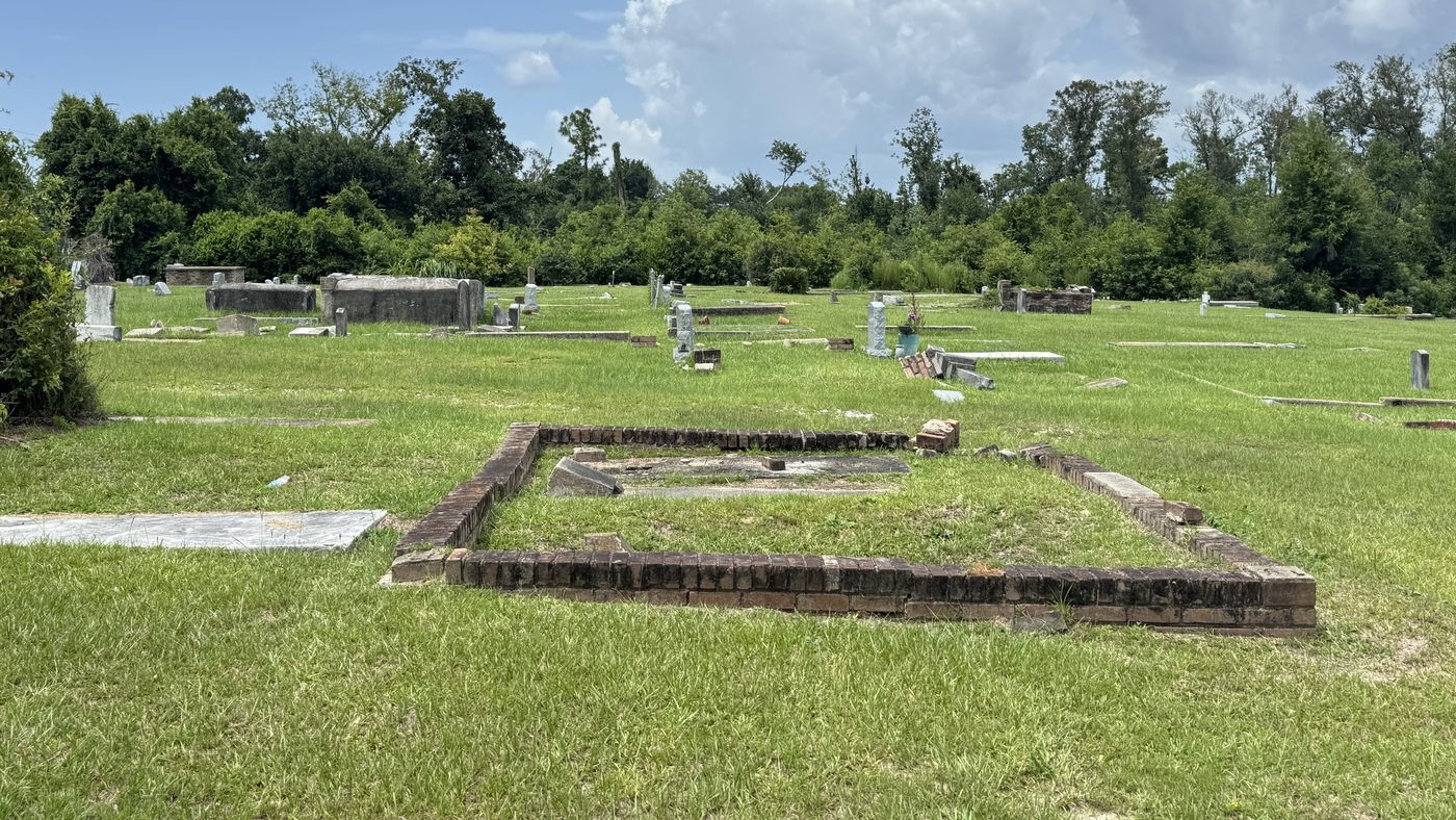 Old graves and brick-bordered plots on the open green of Saint Paul Cemetery, ringed by trees.