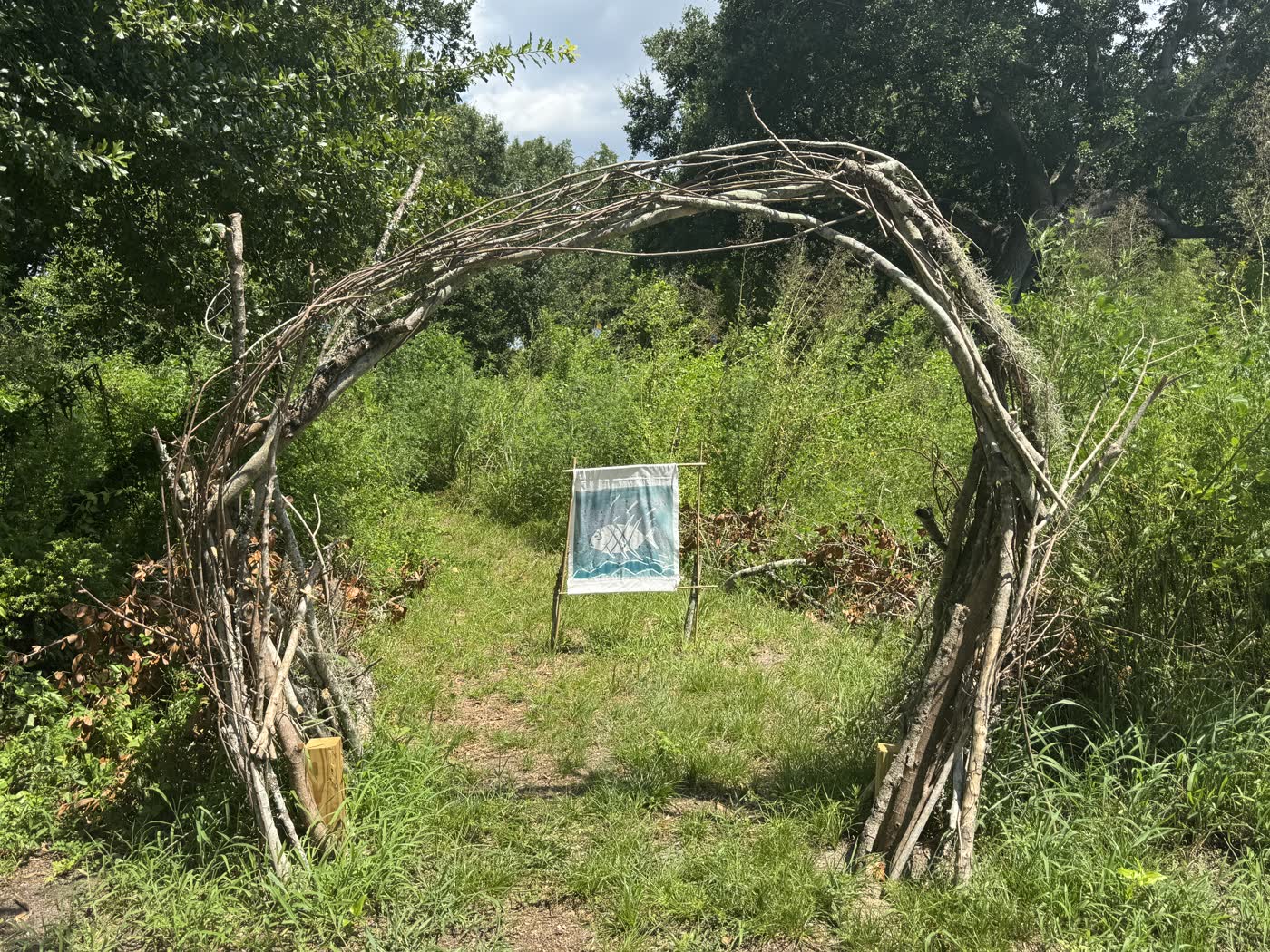 Archway woven from gathered branches at Sawmill Landing Park framing a hand-painted fish banner made by Coast Ephemeral Project students