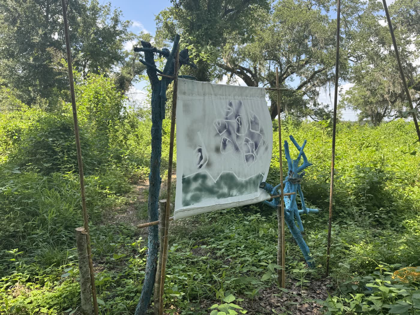 Hand-painted banner hung on a frame of blue-painted branches and bamboo along a Coast Ephemeral Project trail at Sawmill Landing Park