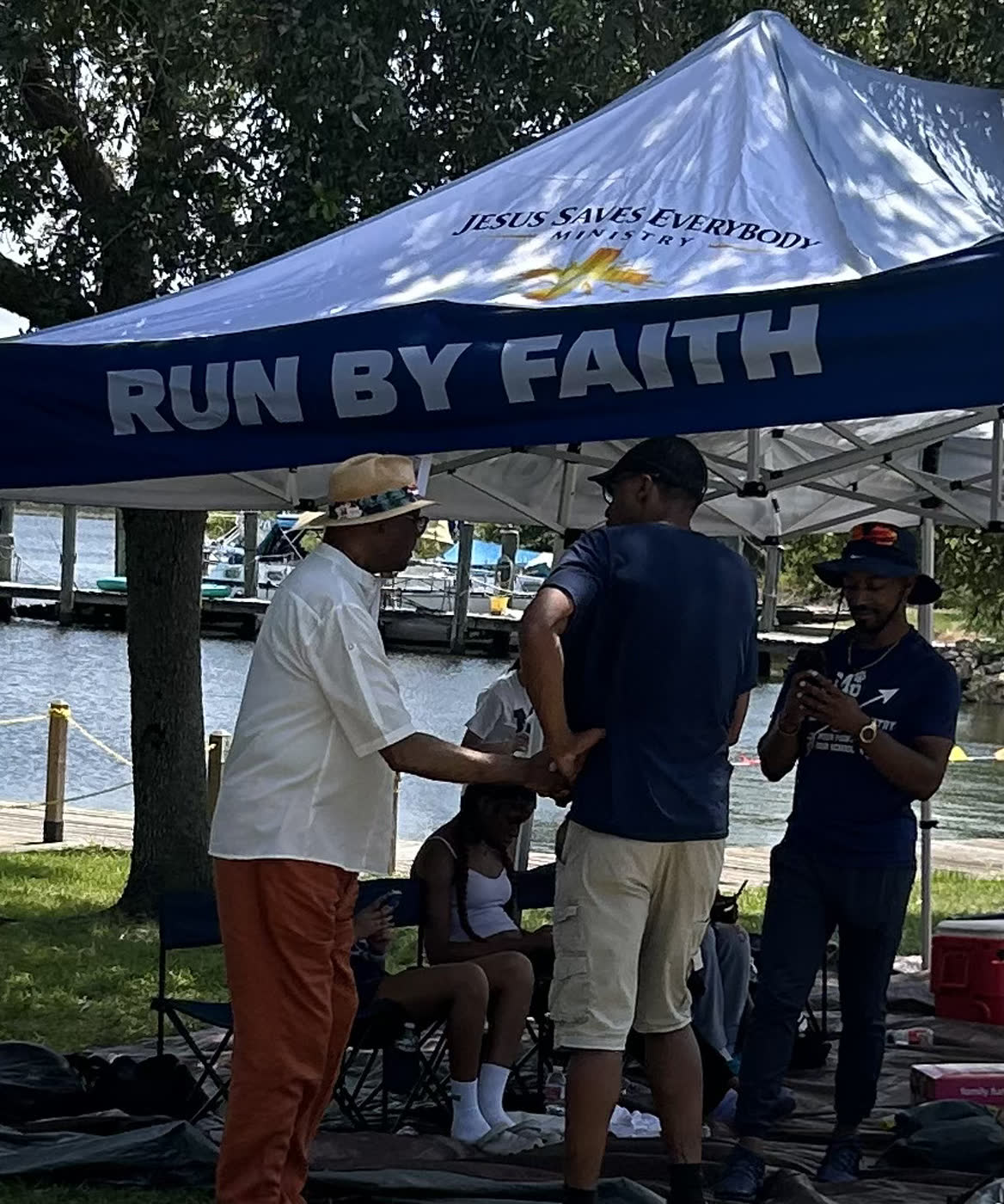 Mayor Billy Knight Sr. greets a resident under a ministry tent at the Faith Fest Fishing Rodeo, with the Escatawpa River and boat slips behind them