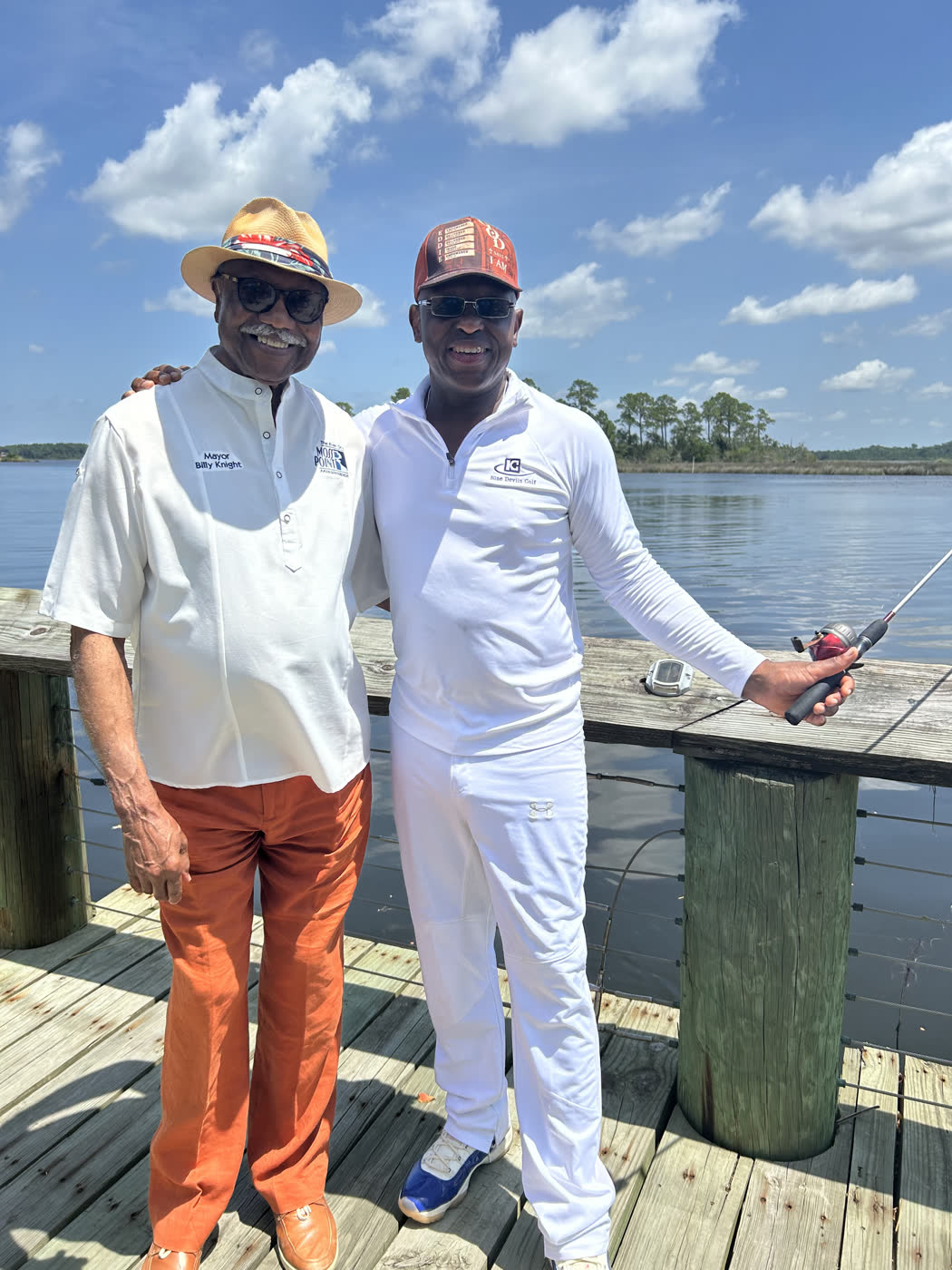 Mayor Billy Knight Sr. stands on the pier with an angler holding a rod, the Escatawpa River behind them, at the Faith Fest Fishing Rodeo